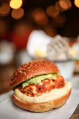 A vertical photo of a crispy chicken sandwich with guacamole on a sesame bun served in a cozy restaurant with warm bokeh lighting, soft focus.