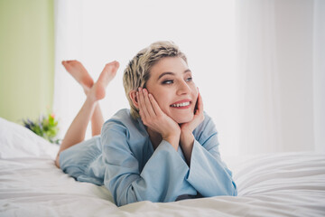 Young woman in blue sleepwear relaxing on a bed and looking cheerful during a bright morning