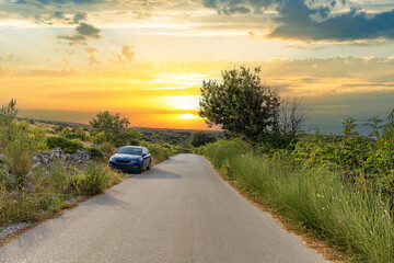 A blue car is parked beside a quiet country road as the sun sets over a lush green landscape, bathing the scene in golden evening light.