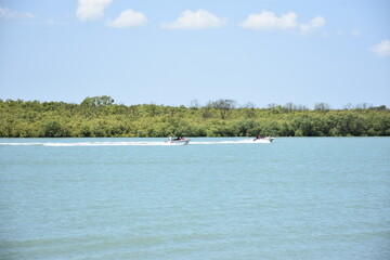 A peaceful view of the Bakkhali River, where a traditional wooden boat glides across calm blue waters. Lush green foliage surrounds the river, creating a serene natural scene.
