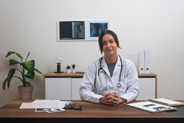 Fototapeta premium Virtual Care Professional: A female doctor, at her desk with patient records, makes eye contact with the camera in an online medical consultation. 