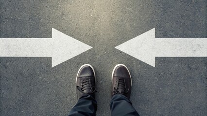 Fototapeta premium Person standing on asphalt road between two opposite arrows. Decision making and direction choice concept.