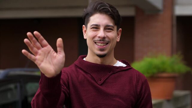 A cheerful young man looks at the camera and waves with a friendly smile, as if meeting a friend or saying goodbye in a warm way
