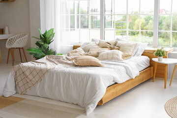 Comfortable bed with lots of soft decorative beige pillows in light sunny bedroom with green plants...