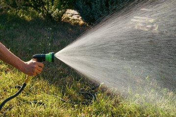 Male hand holding garden hose sprayer watering lawn in backyard on sunny day
