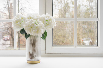 Bouquet of white artificial decorative hydrangea, hortensia in glass vase on windowsill in living...