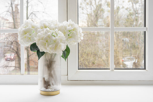 Bouquet of white artificial decorative hydrangea, hortensia in glass vase on windowsill in living room, apartment. Home decor, detail of design interior
