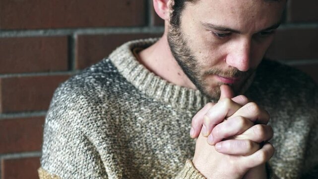 A young man sits near a brick wall with eyes closed, praying silently, hoping to regain balance and clarity after losing his sense of direction in life