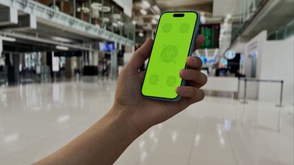 GREEN SCREEN CHROMA KEY A hand holding a smartphone with a green screen in an airport terminal. Blurred travelers and modern architecture in the background