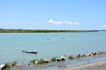 A peaceful view of the Bakkhali River, where a traditional wooden boat glides across calm blue waters. Lush green foliage surrounds the river, creating a serene natural scene.
