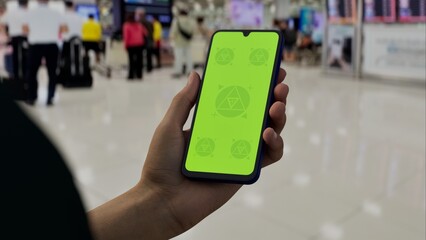 GREEN SCREEN CHROMA KEY A hand holding a smartphone with a green screen in an airport terminal. Blurred travelers and modern architecture in the background