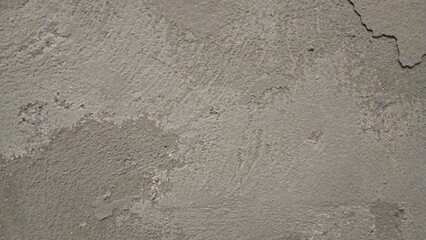 A close-up of a rough, worn-out concrete wall with a gritty texture, featuring cracks and crevices. The color is a dull, muted gray with subtle variations in shade, showing off the uneven surface.