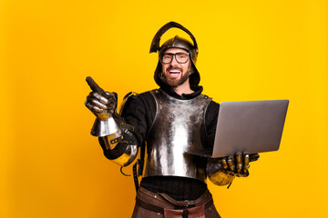 Young man in knight's armor with a laptop and pointing against a yellow backdrop