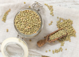 Glass jar full of raw green lentils with a wooden scoop on white table top view. Dried legumes
