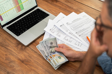 Obraz premium Thoughtful man entrepreneur holds dollar bills checking income sources in digital table on laptop preparing for filling tax forms correctly at workplace