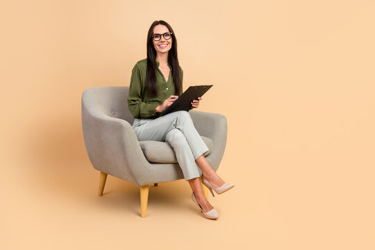 Confident businesswoman with clipboard sitting on beige background, showcasing professionalism and poised demeanor in modern office attire