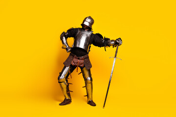 Young man dressed as a medieval knight posing against a vibrant yellow background in full armor...