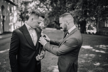 Black and white image of a groom being helped with his boutonniere by a groomsman outside before a...