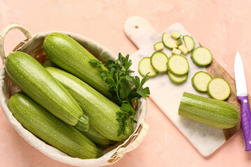 Wicker basket with fresh green zucchini and board of slices on pink background
