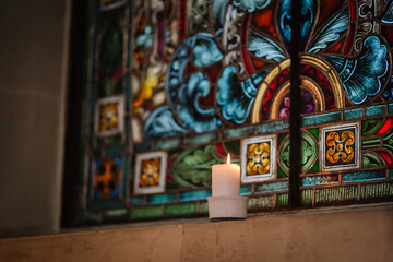A lit white candle glows in front of a colorful stained glass window inside a church, creating a peaceful and spiritual atmosphere..