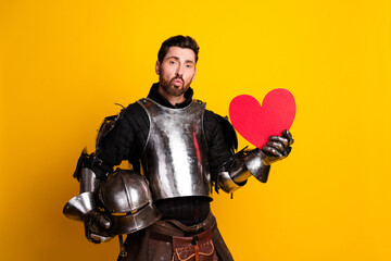 Handsome man in medieval knight armor holding a red heart symbol on yellow background
