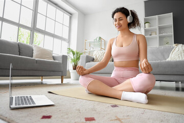 Sporty African-American woman with laptop and headphones meditating on yoga mat at home
