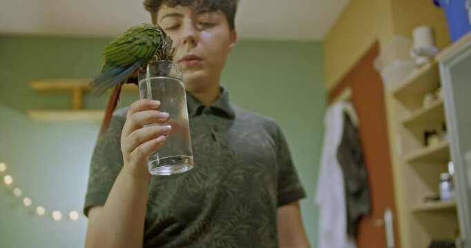A young girl holds a glass of water for her pet green parrot to drink. The parrot perches on the glass and drinks. They are at home.