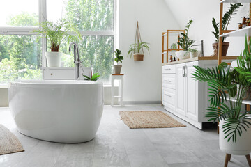 Interior of light bathroom with houseplants, table, bathtub and chest of drawers near window