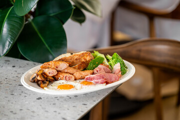 A hearty breakfast plate showcases fried eggs, crispy bacon, sausages, mushrooms, and fresh greens, presented on a white plate, set against a backdrop of soft greenery.
