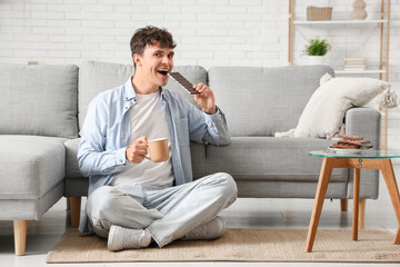 Fototapeta premium Handsome young man with sweet chocolate bar and cup of coffee sitting on carpet in living room