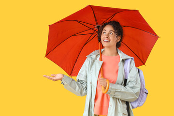 Happy young African-American woman in raincoat with umbrella on yellow background