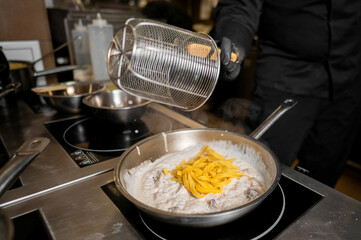 A chef in black gloves prepares a creamy pasta dish in a stainless steel pan, adding fresh pasta to the sauce while steam rises, showcasing culinary artistry in a professional kitchen.