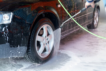 Washing a black car with water and soap in a car washing station during daylight