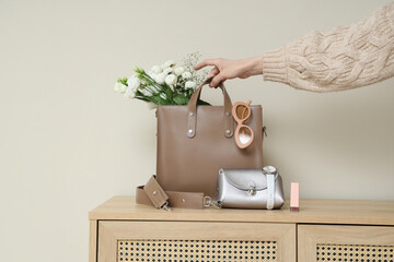 Young woman taking stylish bag with flowers from commode near light wall