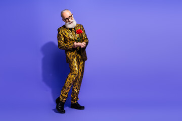 Elegant mature man in an extravagant gold patterned suit holding a red rose with a confident pose against a vibrant violet background