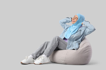 Beautiful young Muslim woman in hijab sitting on beanbag chair against grey background