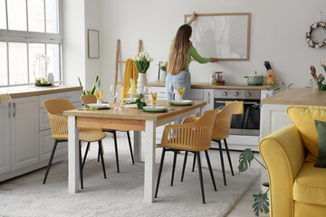 Young woman hanging blank frame on light wall in kitchen decorated for Easter