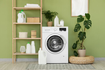 Modern washing machine with shelving unit, houseplants and detergents near green wall in laundry room
