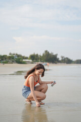 Naklejka premium woman in bright top and denim shorts joyfully crouches on serene beach, holding sunglasses, with trees and calm water in background