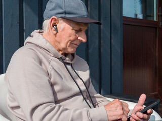 Senior man relaxes outdoors using smartphone with wireless earbuds in sunny garden. Senior man enjoys calm digital moment. Senior man embraces peaceful technology-driven lifestyle. 