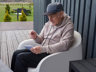 Elderly man sits comfortably in modern white chair, wearing baseball cap and writing notes in notebook. Elderly man enjoys peaceful moment. Elderly man embraces creative relaxation. 