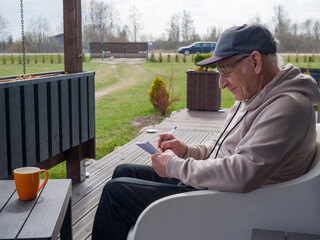Elderly man sits comfortably in modern white chair, wearing baseball cap and writing notes in notebook. Elderly man enjoys peaceful moment. Elderly man embraces creative relaxation. 
