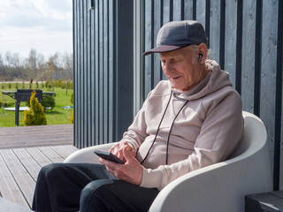 Senior man sits comfortably outdoors in hoodie and cap, listening to music through wireless earphones. Senior man scrolls smartphone. Senior man enjoys peaceful tech-savvy leisure time. 