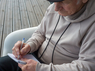 Concentrated senior writing notes in notebook while sitting on white chair near wooden wall, capturing moment of focused reflection and quiet contemplation during retirement leisure