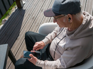 Senior man wearing baseball cap and hoodie enjoying music through wireless earbuds, standing against dark wooden background, experiencing peaceful moment of personal audio connection