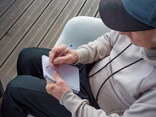 Elderly man sits quietly on wooden porch, writing thoughtful notes in personal notebook. Elderly man enjoys peaceful garden view. Elderly man embraces calm outdoor moment. 