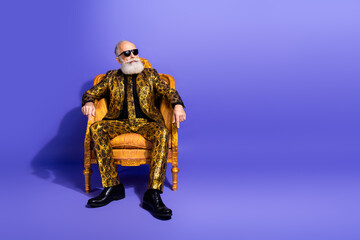 Trendy senior man with white beard seated in ornate chair against vibrant purple background showing elegance and eccentric fashion style