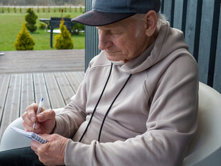 Elderly man in newsboy cap sits in white armchair on terrace, writing notes beside cup of tea. Elderly man enjoys calm moment. Elderly man focuses on peaceful creative routine.
