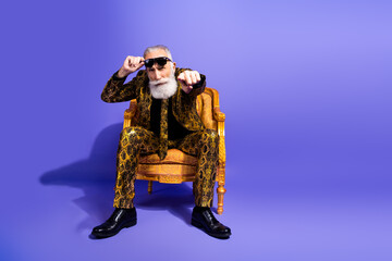 Stylish senior man in a golden patterned suit posing confidently on a purple background, showcasing elegance with a hint of eccentricity