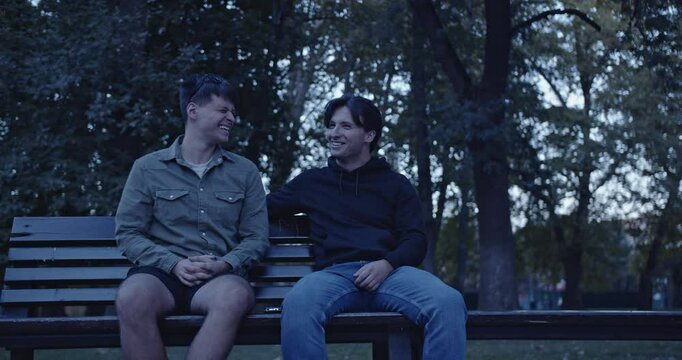 Two young men sit on a park bench, talking and laughing together. They enjoy each other's company on a warm summer evening.